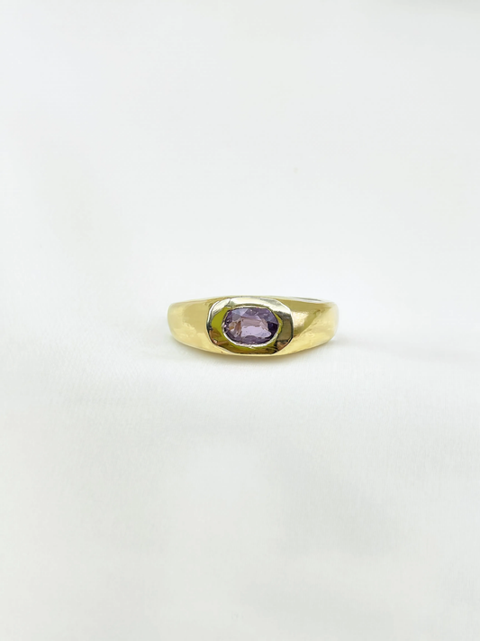 A gold signet-style ring with a bezel-set oval purple sapphire, photographed on a plain white background.