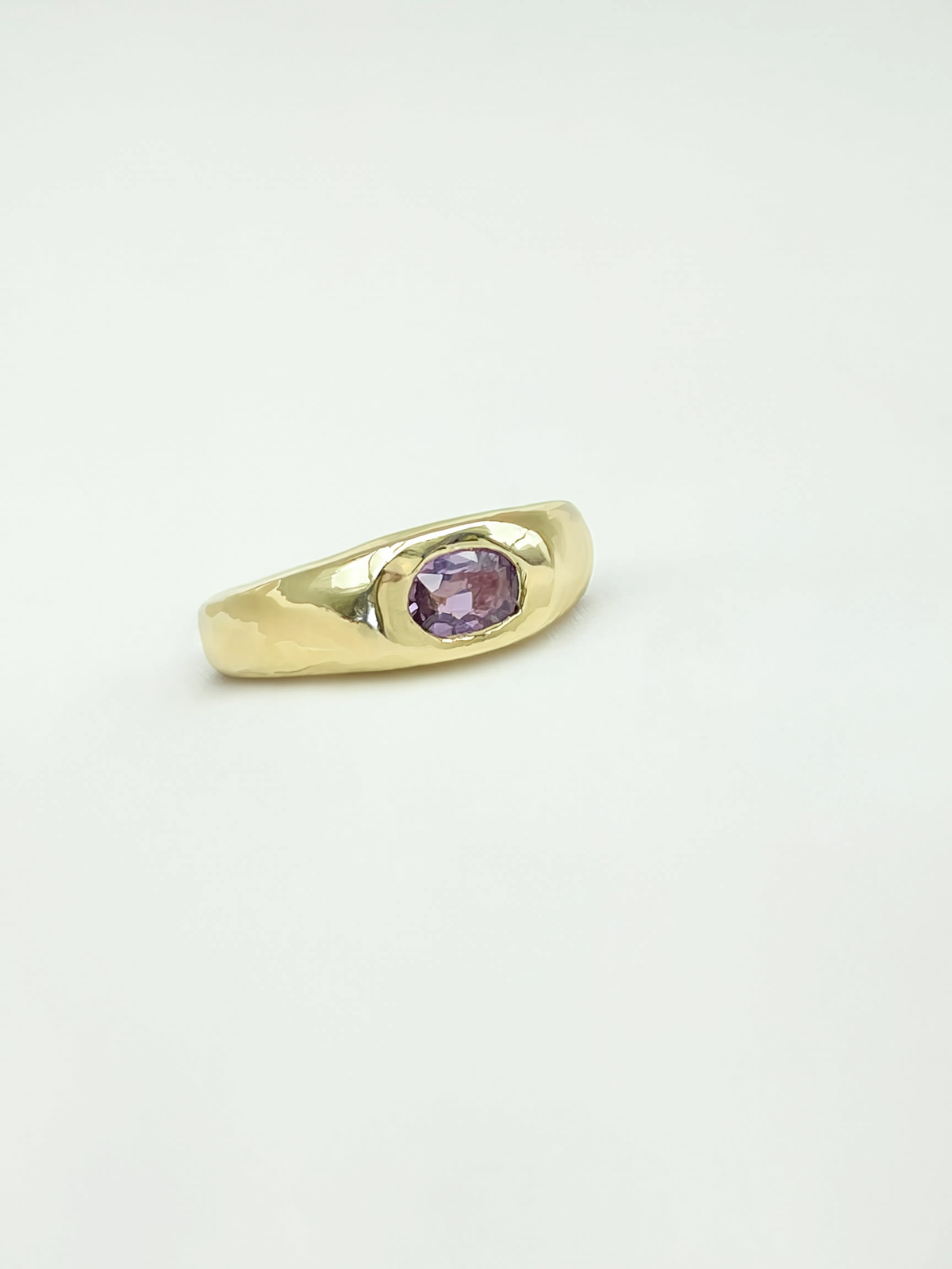 A gold signet-style ring with a small oval purple sapphire, photographed on a plain white background.