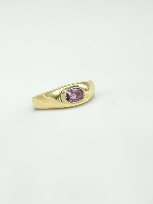 A gold signet-style ring with a small oval purple sapphire, photographed on a plain white background.