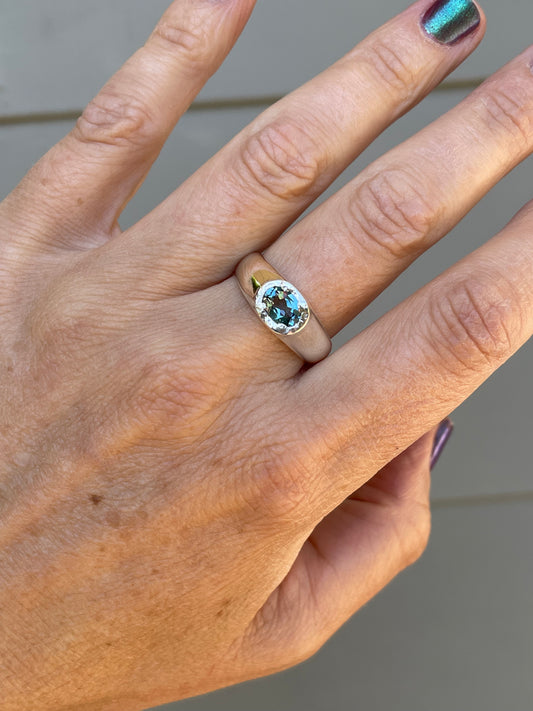 A close-up of a hand wearing a silver ring with a round-cut light blue gemstone, set against a neutral gray background.