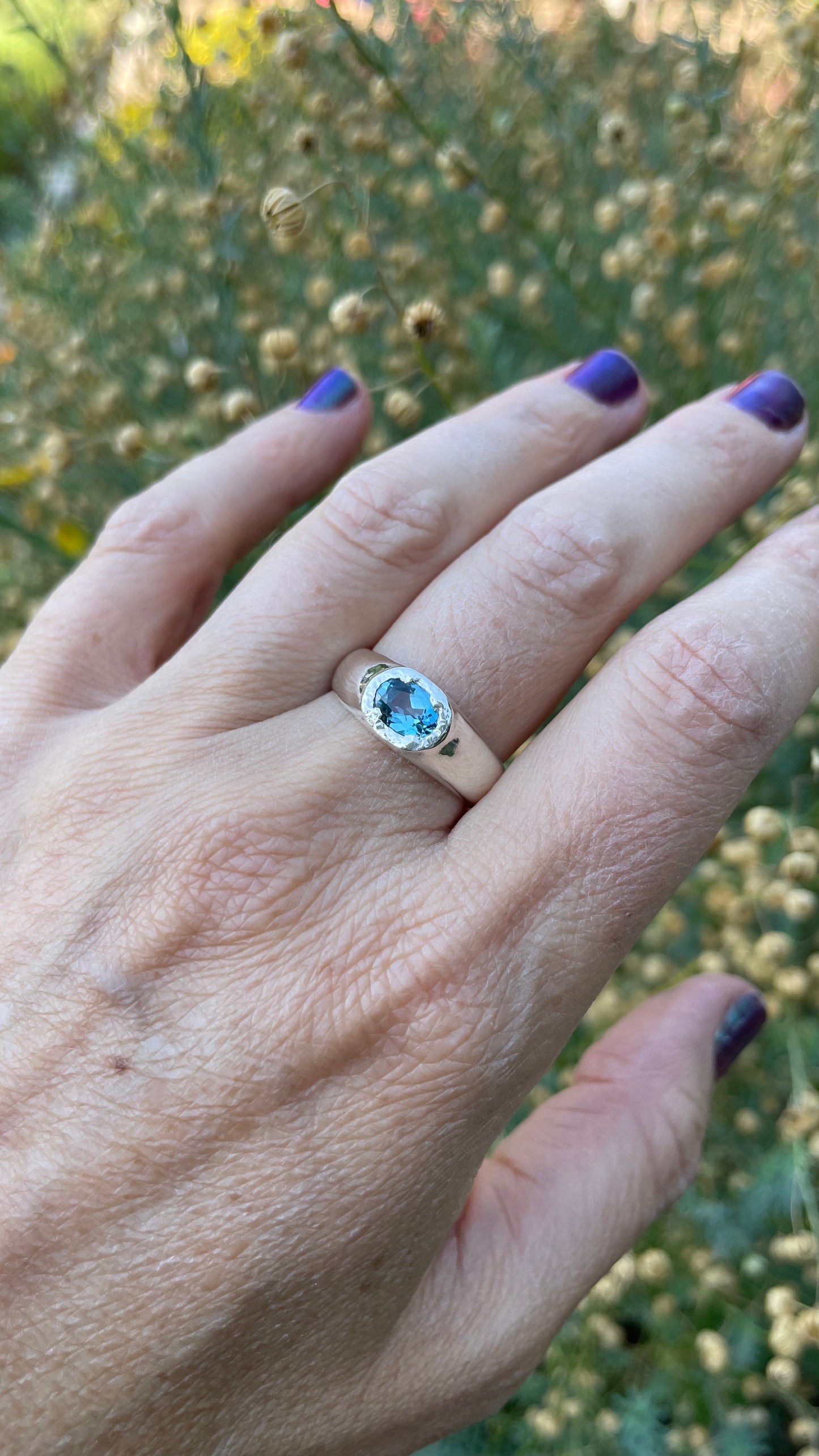 A silver ring with a round-cut light blue gemstone worn on a person's ring finger, shown against a blurred background of green foliage and small yellow flowers.