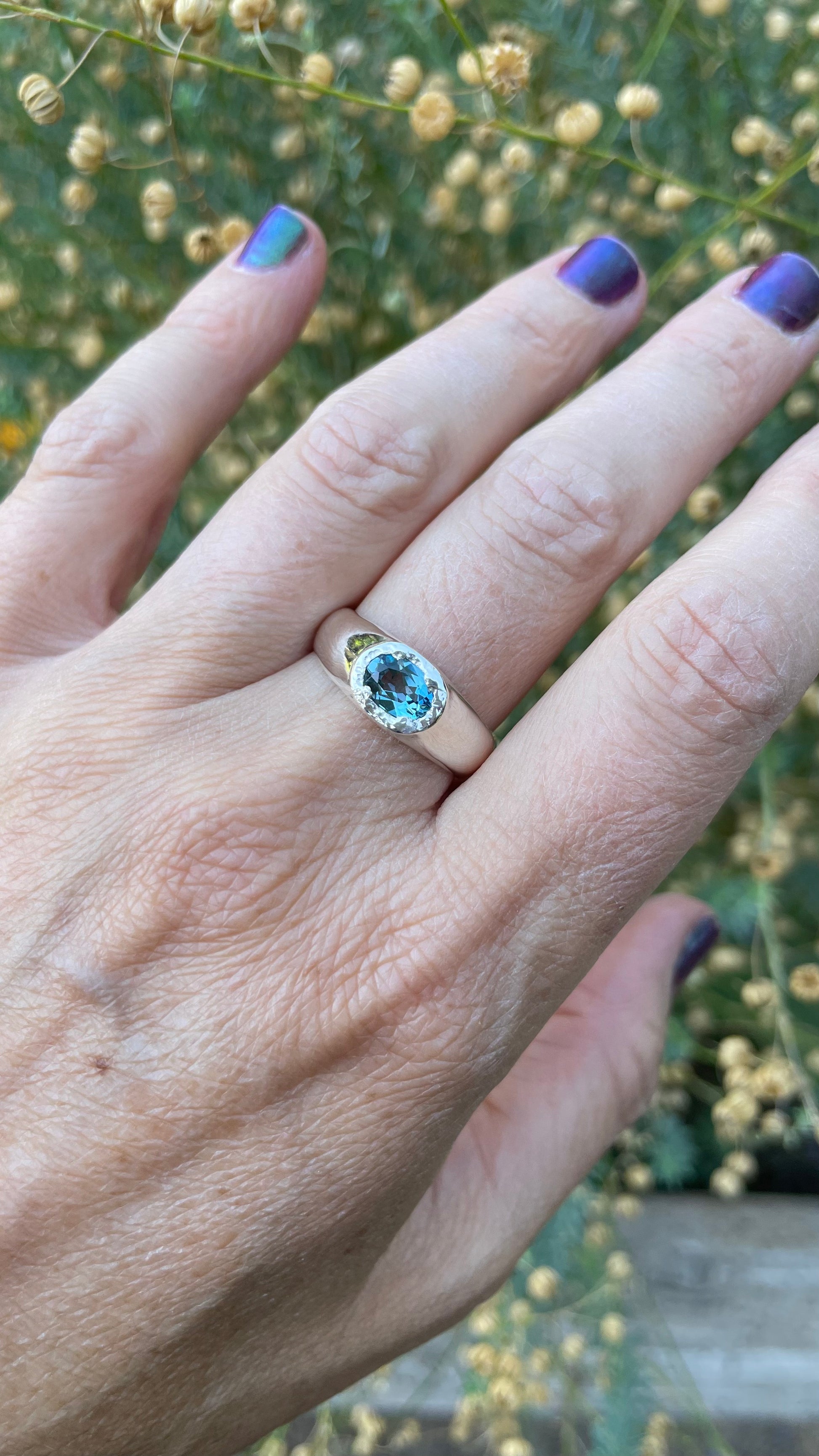 A close-up of a hand wearing a silver ring with oval deep London blue topaz, set against a soft-focus background of green foliage and small yellow flowers.