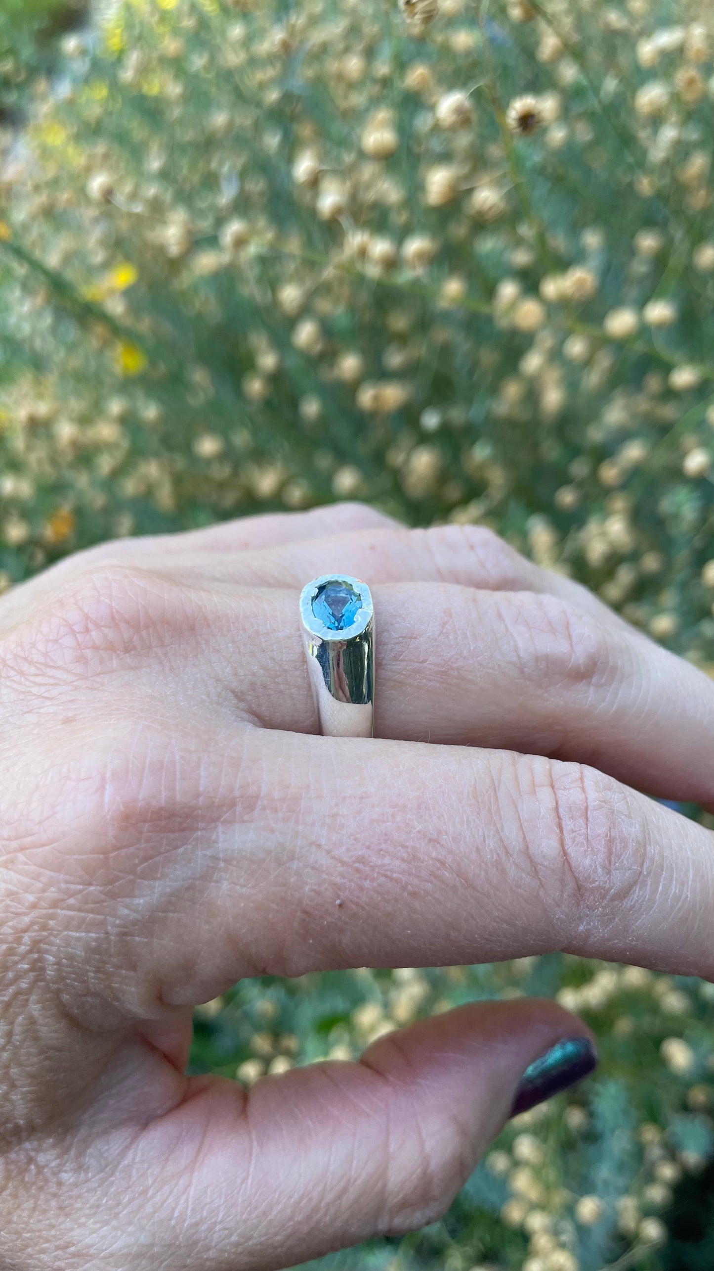 A silver ring with a round blue gemstone worn on a person's finger, photographed outdoors against a blurred background of green foliage and small beige flowers.