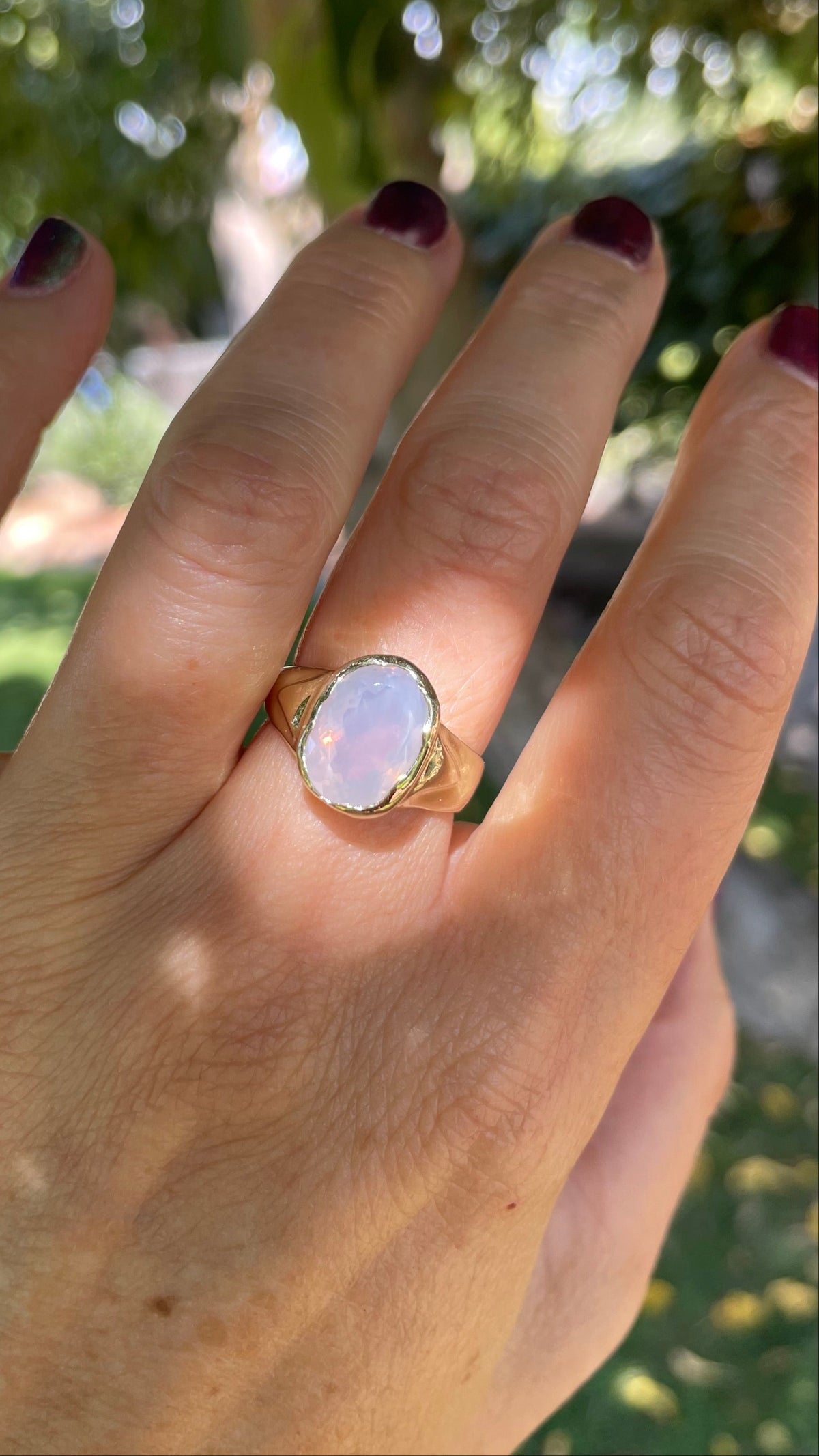 A gold ring with an oval lavender quartz gemstone worn on a person's ring finger, shown against a blurred outdoor background of green foliage.