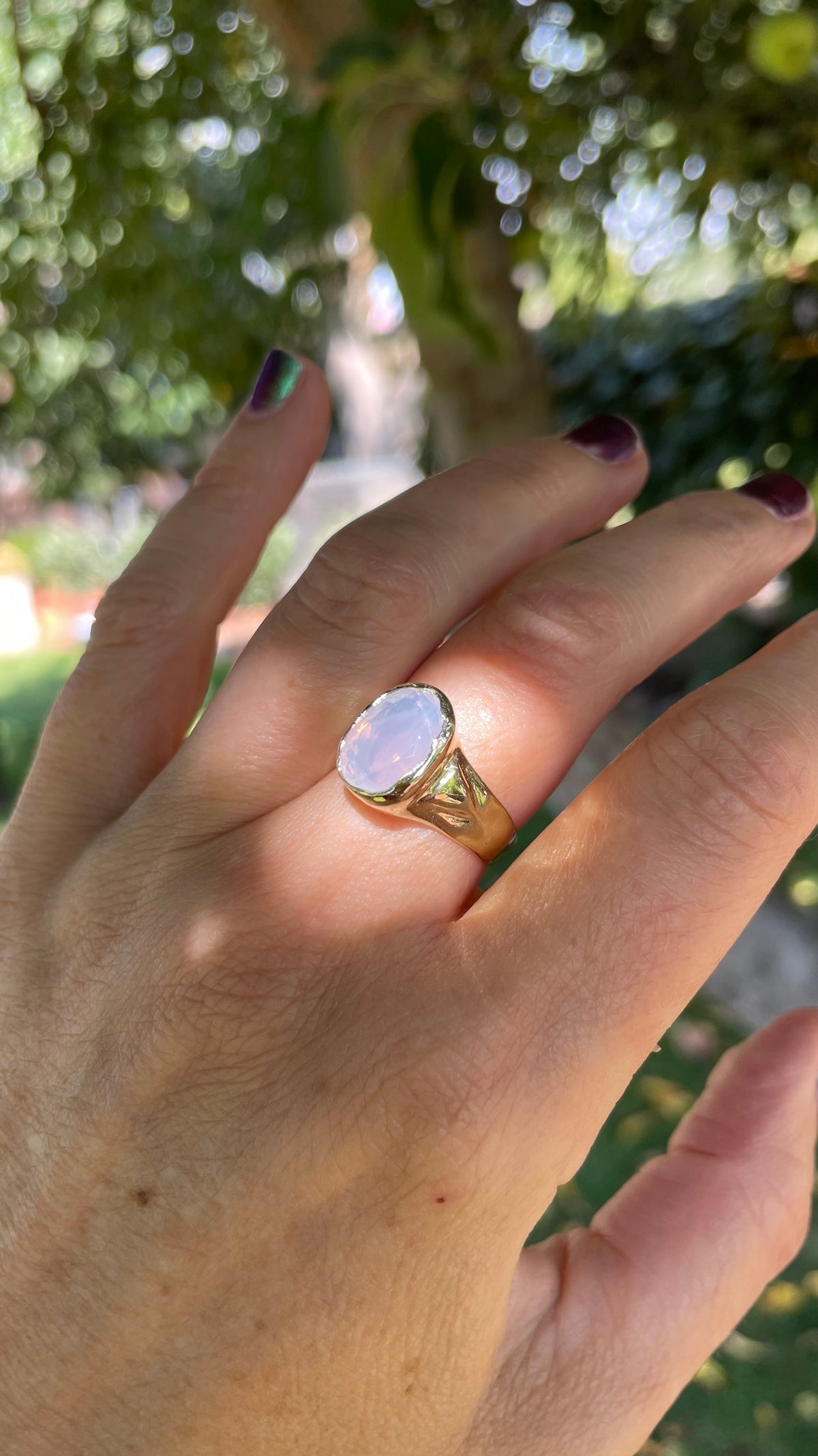 A gold signet-style ring with a large oval pale pink stone worn on a person's ring finger, photographed outdoors with a blurred green garden background.