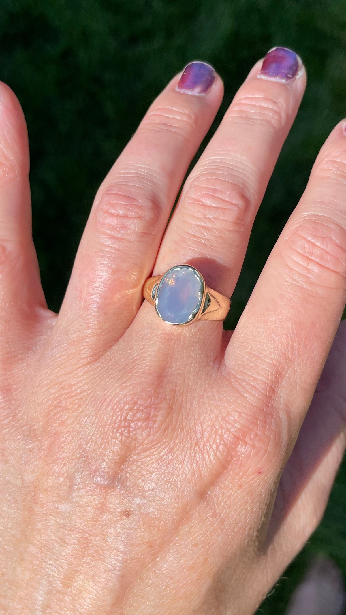A gold signet ring with an oval lavender blue-pink stone worn on a person's ring finger, shown against a background of green grass.