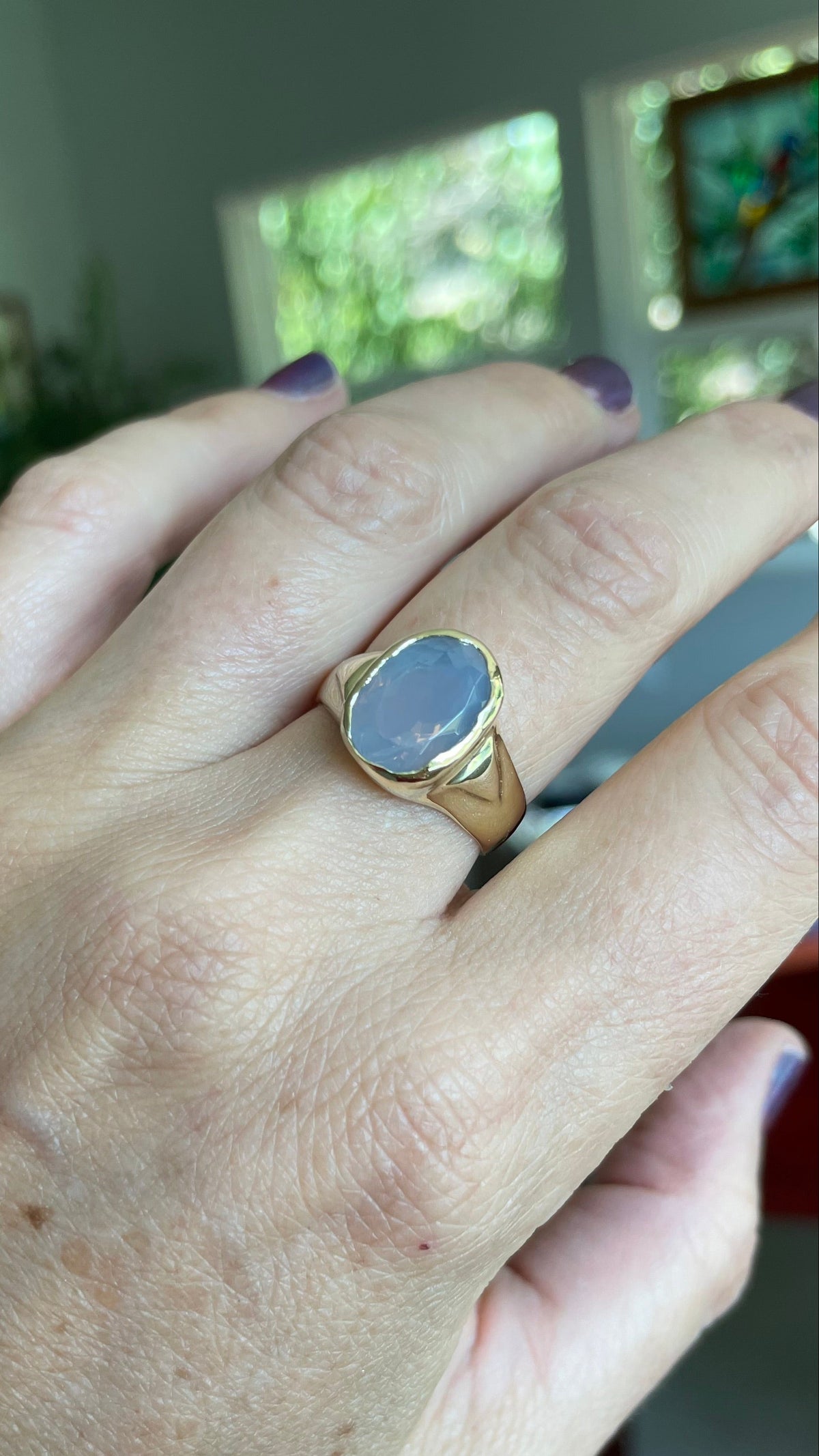 A gold ring with a large oval lavender blue stone worn on a person's hand, shown in a close-up shot with a blurred indoor background.