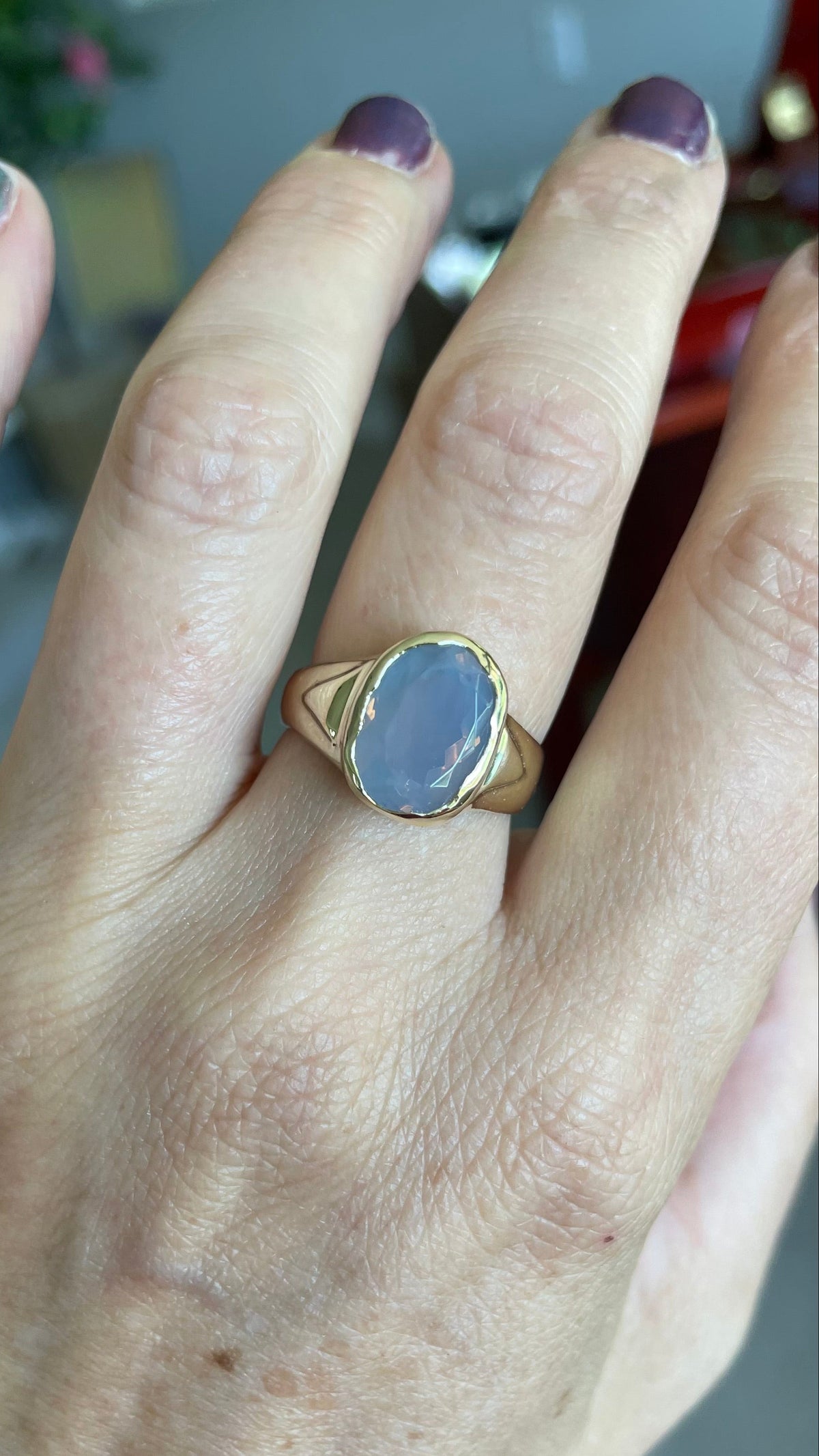A gold signet ring with an oval light blue-lavender quartz stone worn on a person's ring finger, shown in a close-up shot of the hand.
