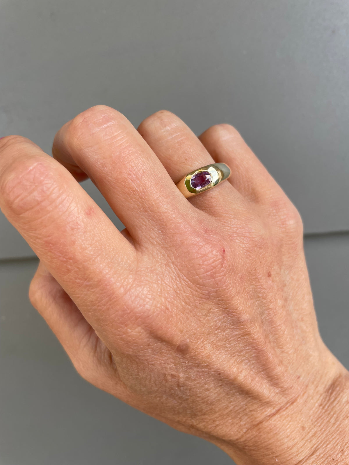 A close-up of a hand wearing a gold signet ring with a round purple gemstone, set against a plain gray background.
