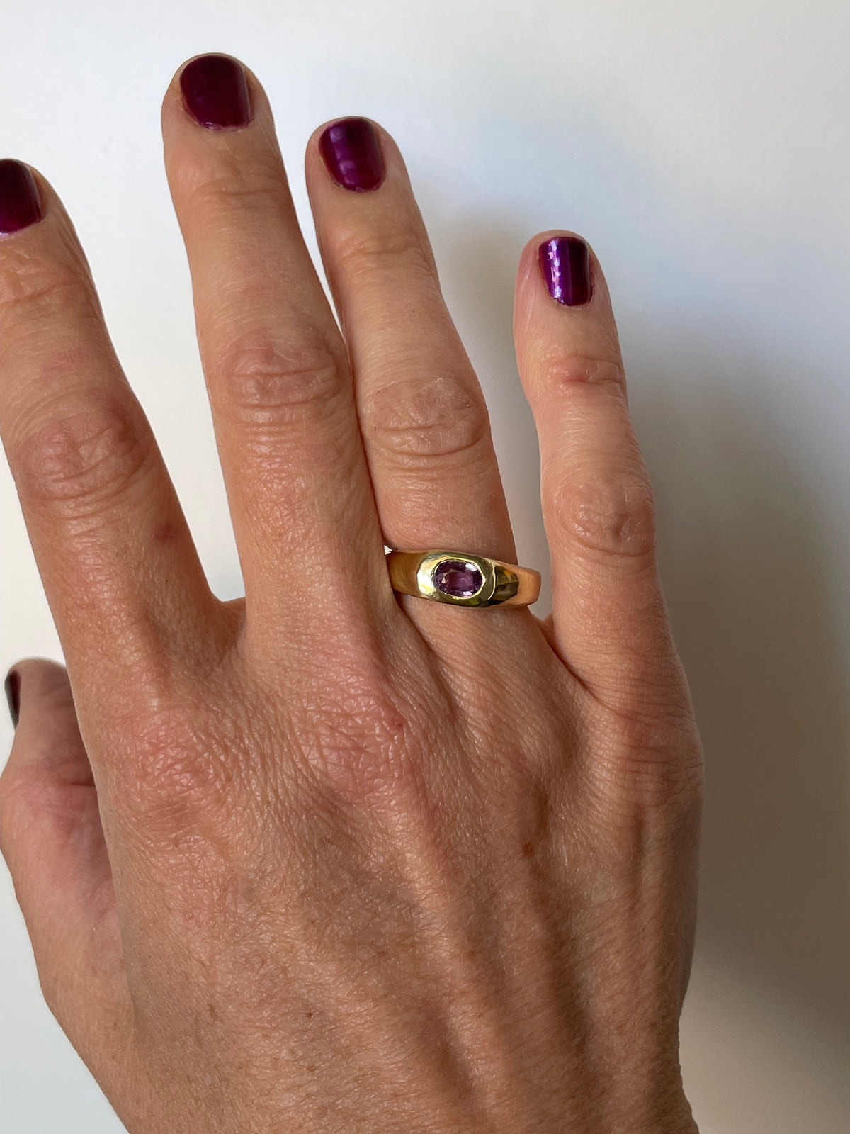 A gold ring with a small oval purple gemstone worn on a person's ring finger, shown against a plain light background.